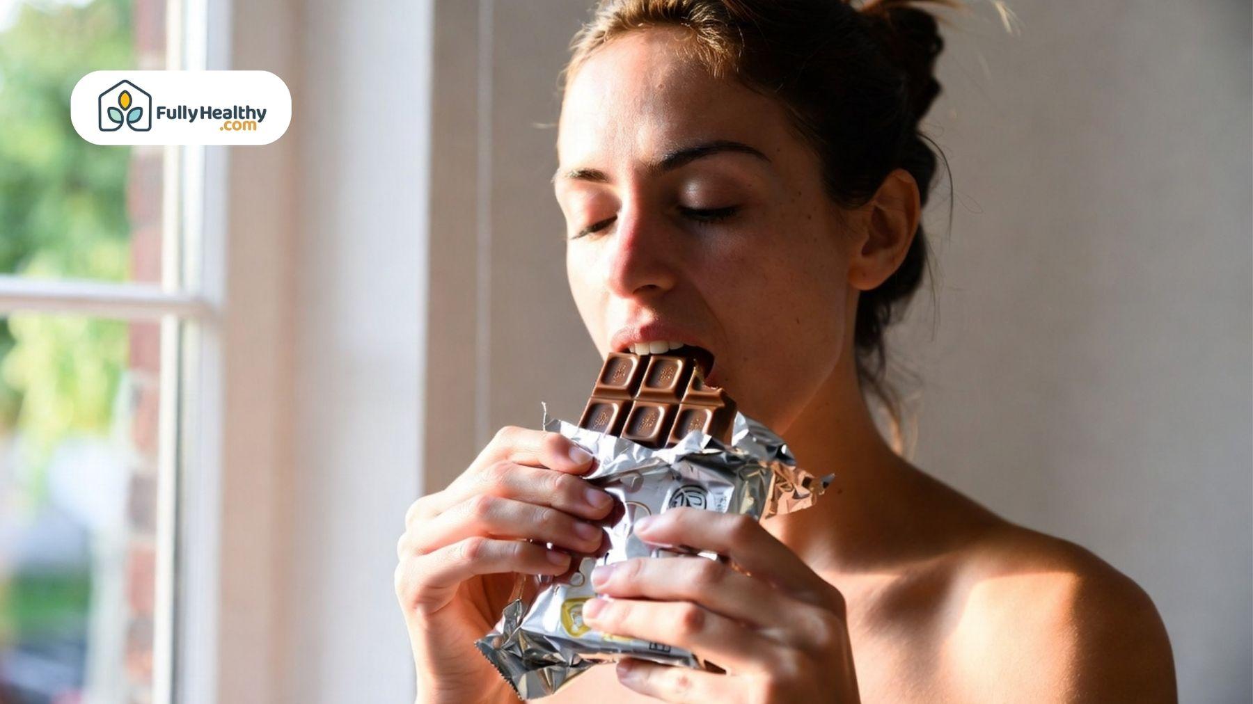 Person taking a bite of a chocolate bar near window light.