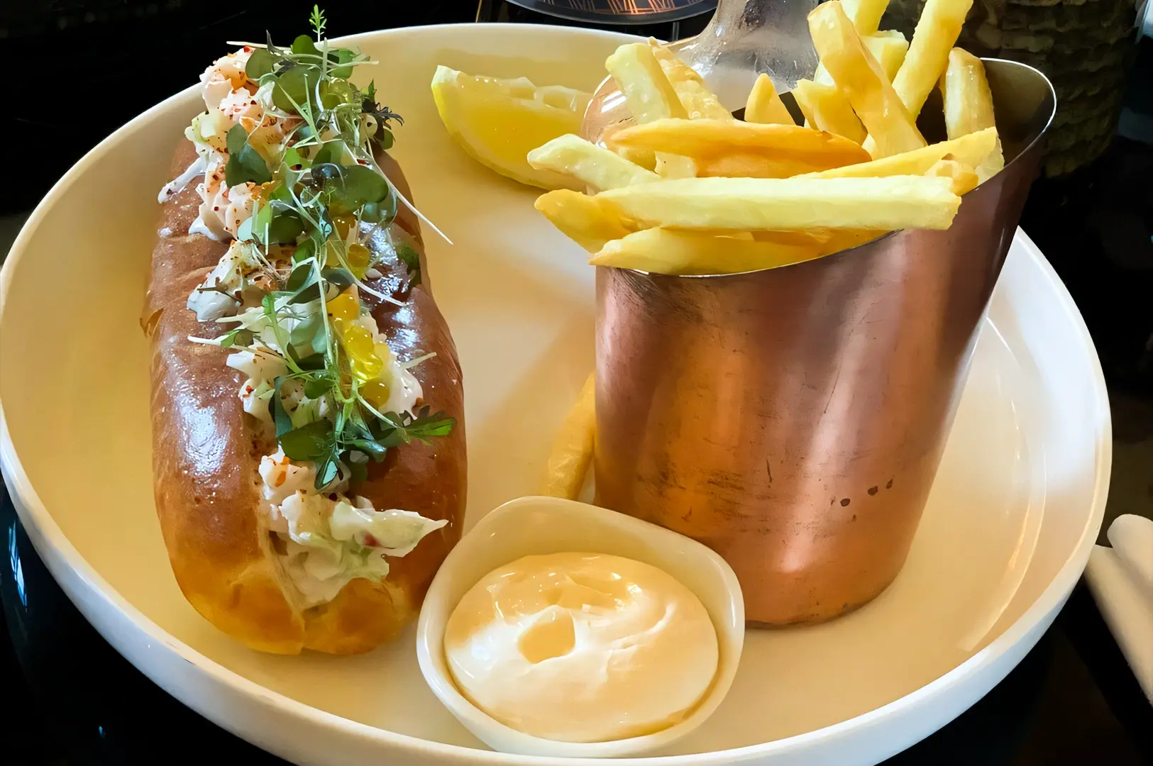 Three‑quarter angle close‑up shot of a lobster roll topped with herbs, served with golden fries in a copper cup and dipping sauce on a white plate inside Atlas Bar Singapore.
