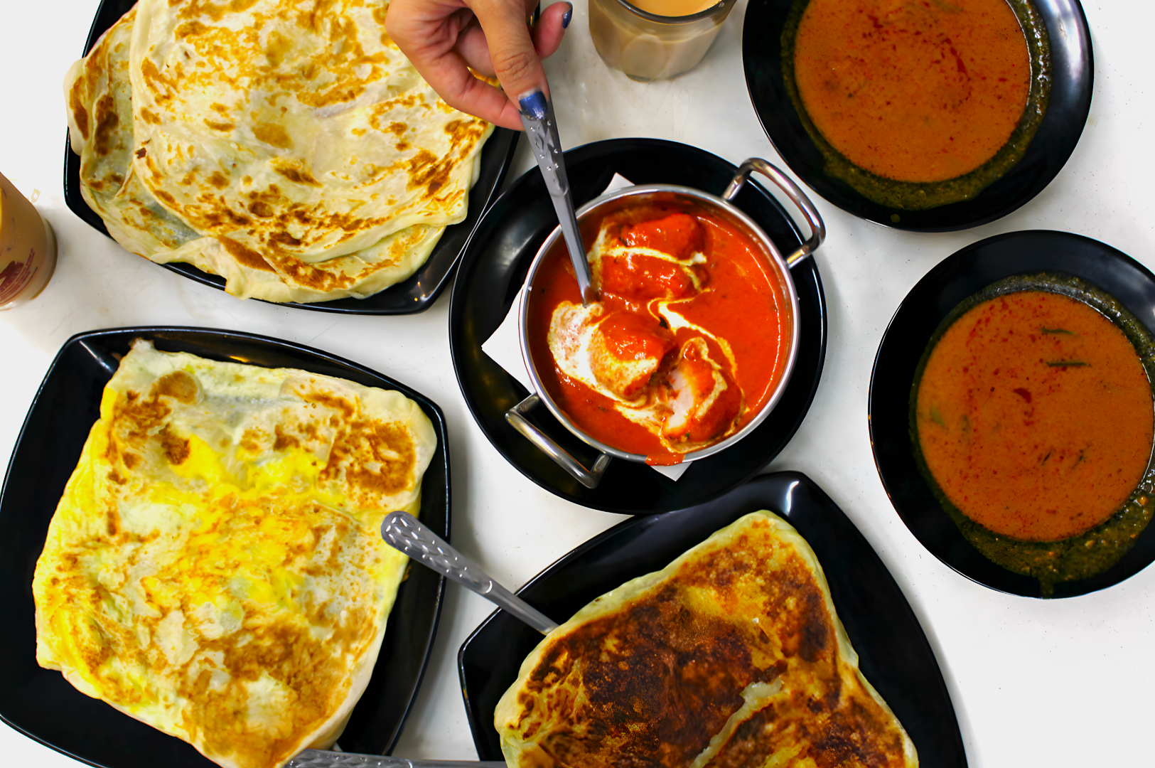 A hand dipping naan into rich, red curry surrounded by various stuffed flatbreads and two bowls of curry on black plates, evoking a warm, hearty meal.