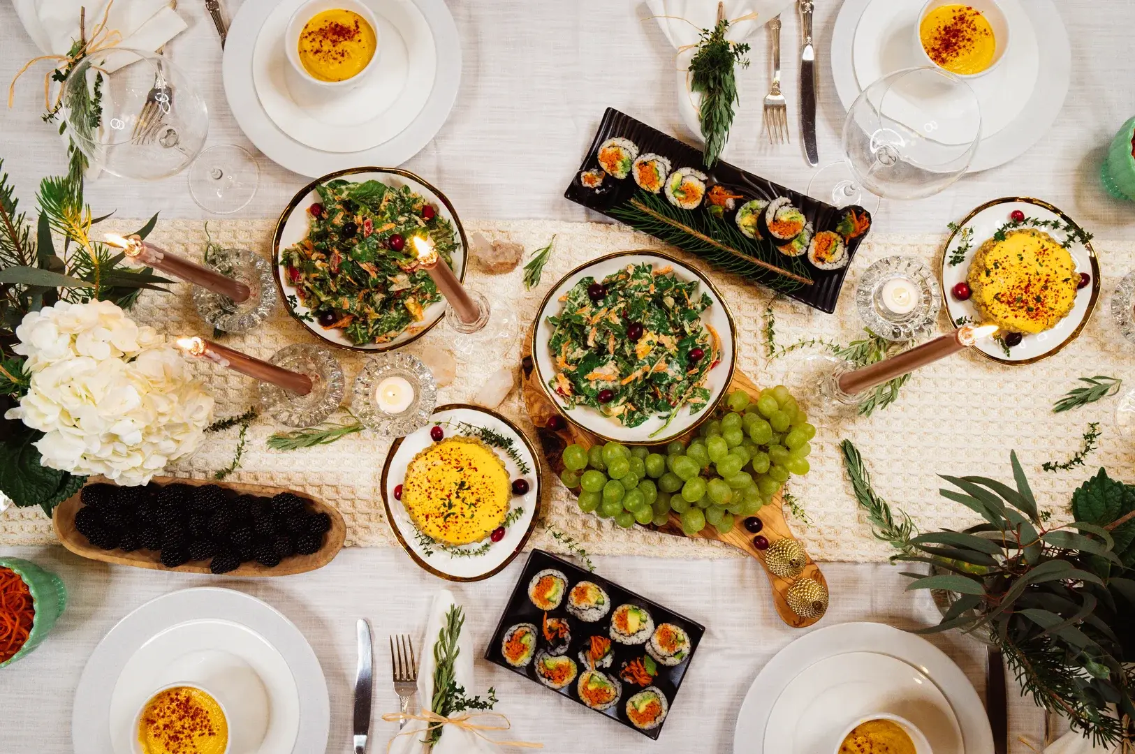 Overhead flat lay shot of an elegant festive dining table spread featuring sushi rolls, leafy salads, grapes, berries, baked custards, candles, and floral decor arranged on a neutral tablecloth for a stylish holiday dinner or gourmet party setting.