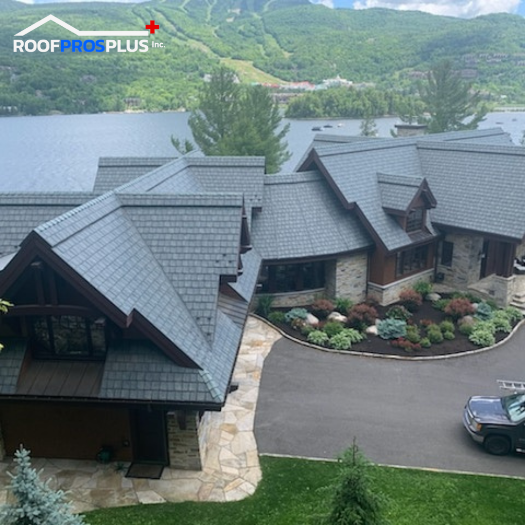 A large, elegant lakeside house with a grey metal roof and landscaped garden. The scenic view includes a tranquil lake and lush green mountains. The logo of Roof Pros Plus is visible in the top left corner of the picture.