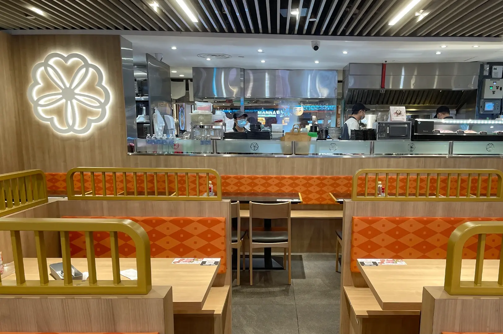 Eye-level straight-on interior shot of a modern casual dining restaurant featuring orange upholstered booth seating, light wood tables, an open stainless-steel kitchen, and warm ambient lighting for SEO-friendly “modern restaurant interior open kitchen booth seating design.