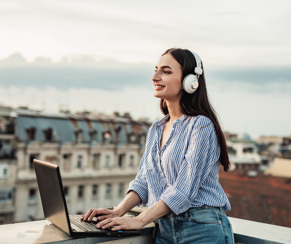 a smiling woman in a button down shirt works on her laptop while wearing headphones on a rooftop