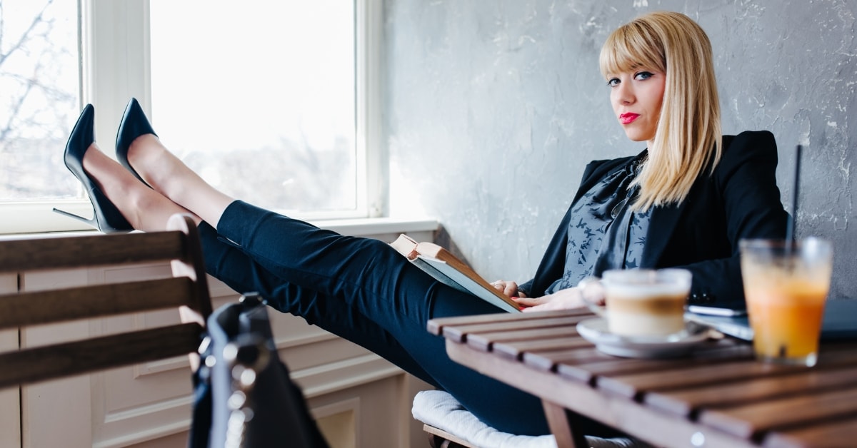 A woman with blonde hair in a black suit sits with her legs crossed on a table, representing an OnlyFans taxing authority presence.