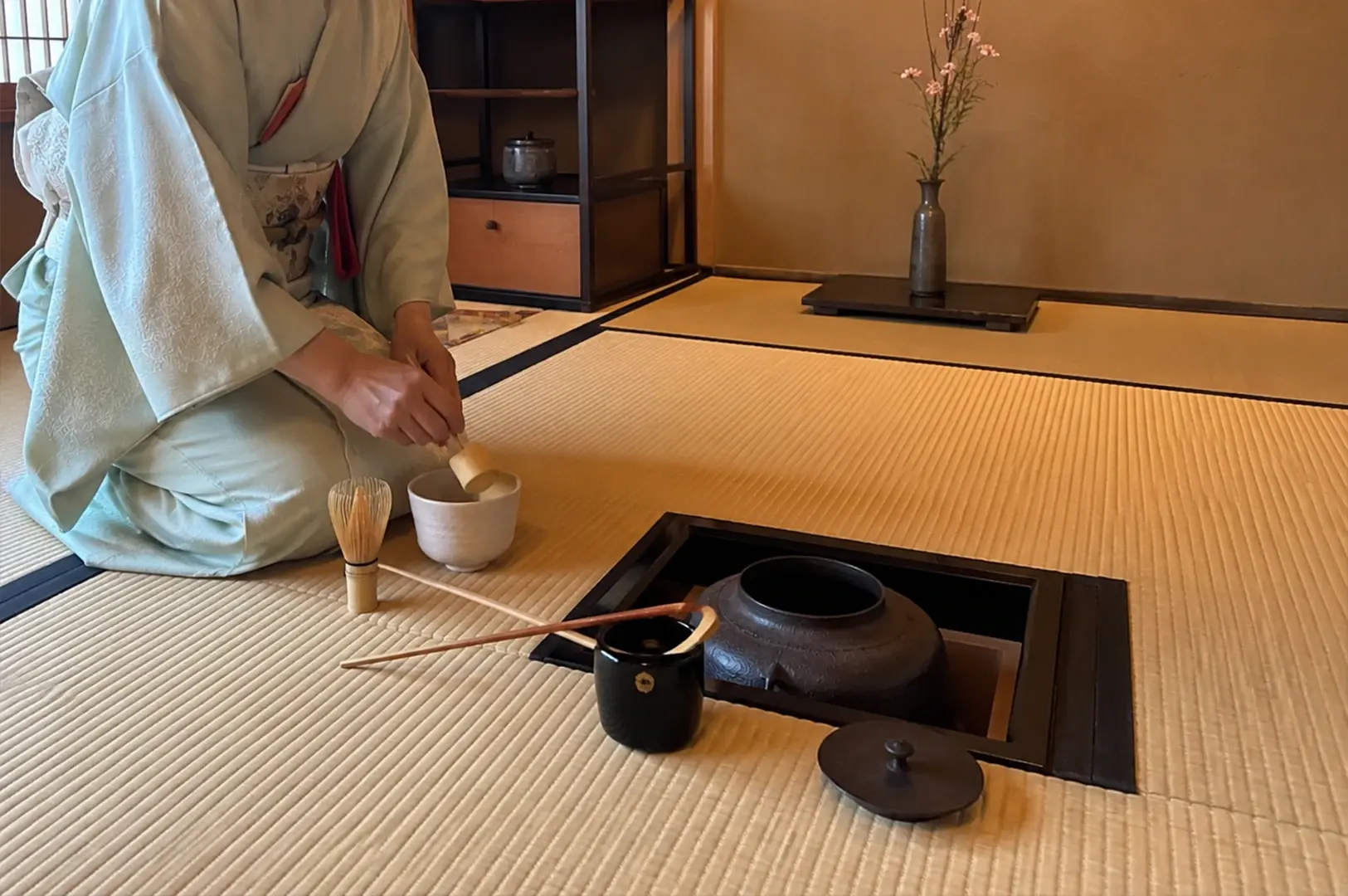 A high-angle view of a tea practitioner dressed in a pale blue kimono kneeling on a tatami mat floor while preparing matcha. The practitioner uses a wooden ladle over a white ceramic bowl, situated next to a square sunken hearth that contains a dark, textured iron kettle with its lid resting nearby. A bamboo whisk, a long bamboo scoop, and a small black ceramic jar are neatly arranged around the hearth, while the background shows a minimalist room with a wooden shelf and a slender vase holding delicate pink flowers.