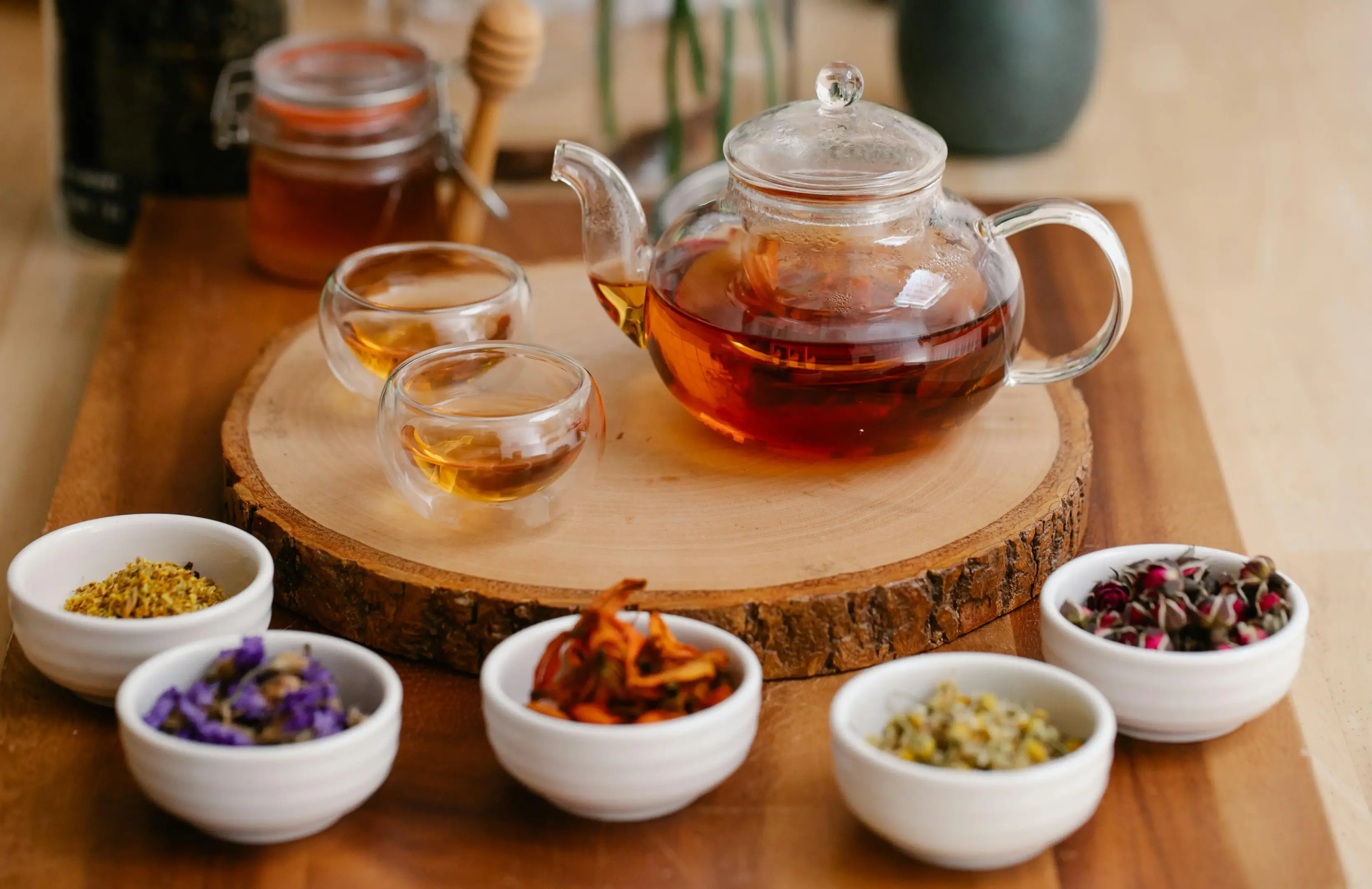 A glass teapot filled with amber-colored tea sits on a wooden platter alongside two small, matching double-walled glass cups. Five small white bowls containing various colorful dried herbs and flowers, such as rosebuds and chamomile, are arranged in front of the platter.