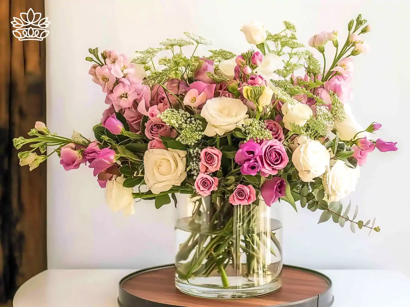 A beautifully arranged bouquet featuring an assortment of white and pink roses, delicate lavender blooms, and lush greenery, elegantly displayed in a clear glass vase on a wooden surface. This floral arrangement exudes charm and sophistication, perfect for enhancing any space. All Flower Arrangements Collection. Fabulous Flowers and Gifts.