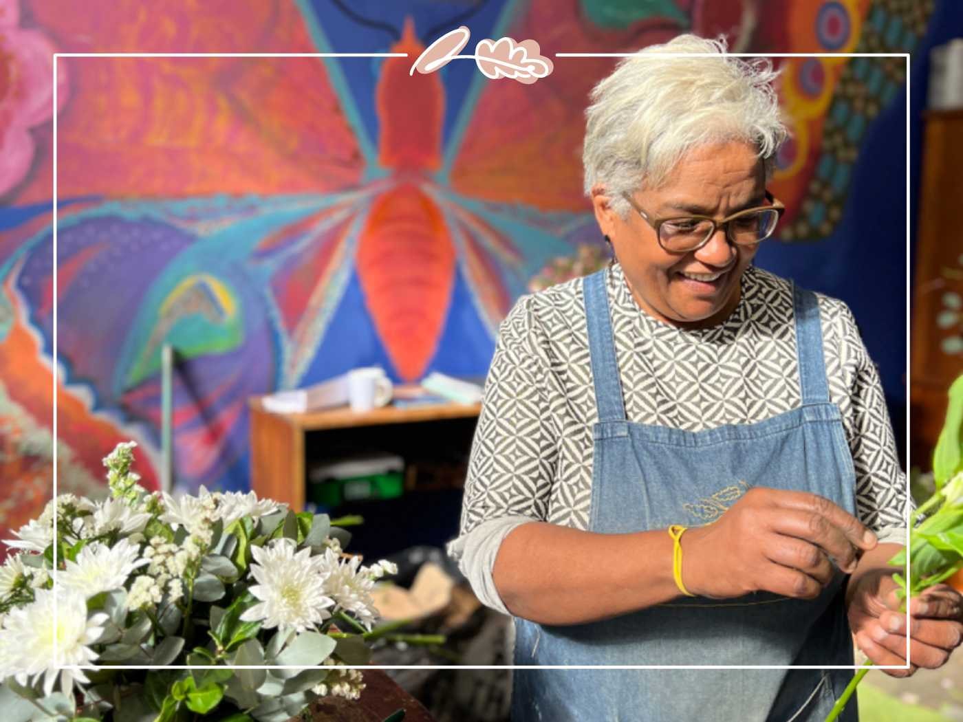 A joyful woman with gray hair wearing glasses and a patterned apron smiles as she works on a floral arrangement in front of a colorful mural. Fabulous Flowers and Gifts.