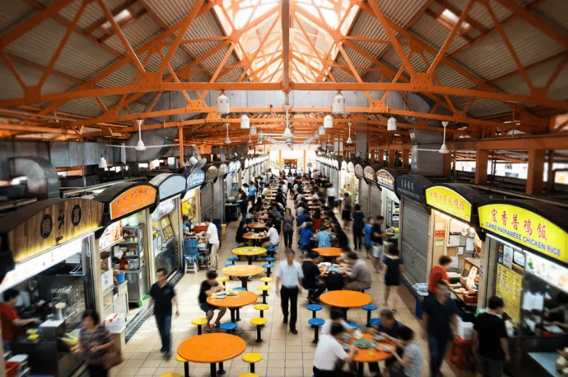 Wide view of the bustling Maxwell Food Centre interior, showing rows of hawker stalls under an orange vaulted ceiling and patrons dining at round orange tables along the central aisle.