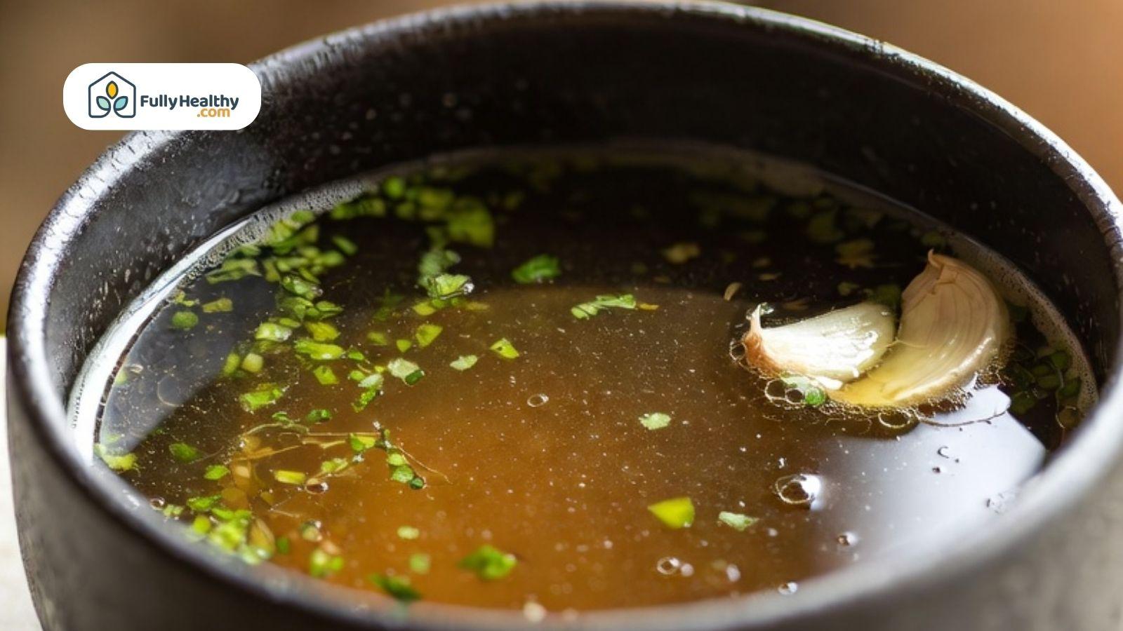 Warm bone broth in a dark bowl with chopped herbs and a clove of garlic on the surface.