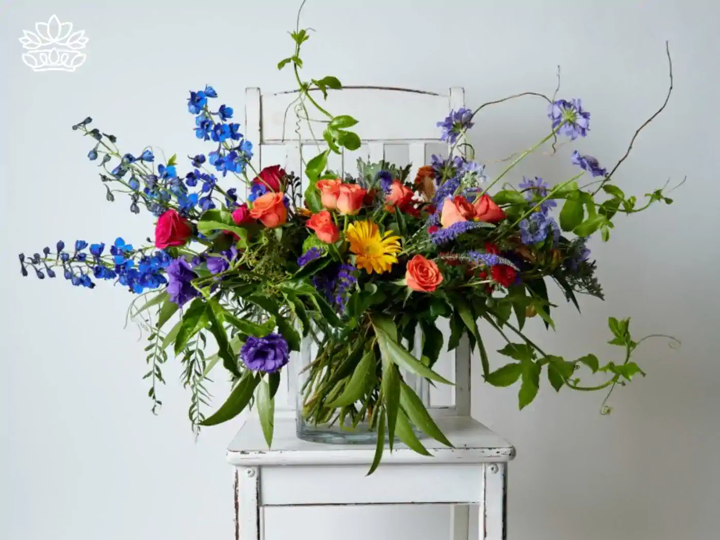 Lush and colorful front desk floral reception desk arrangement in a rustic glass vase, featuring an array of blue delphiniums, vibrant red and orange roses, and wild greenery on a vintage white chair.