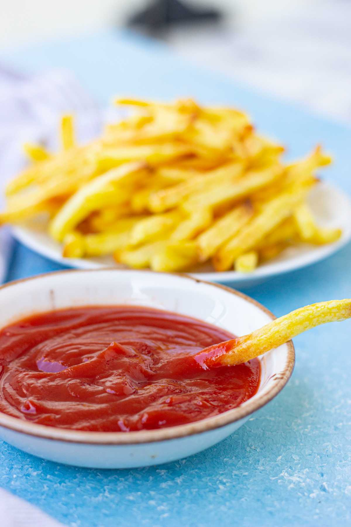 A bowl of ketchup being dipped a piece of fries and a plate of cooked fries at the back.