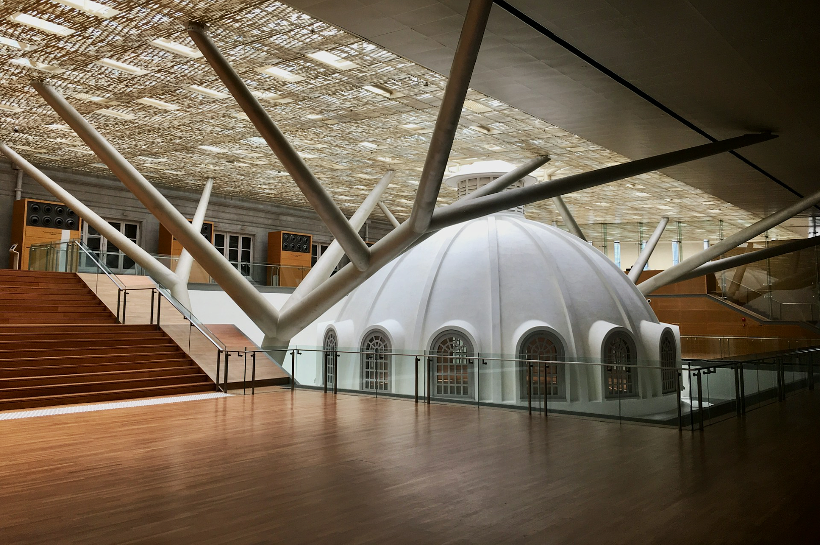 Modern interior with wooden floors, a large white dome, and angled beams. Glass railings surround the dome. The atmosphere is spacious and airy.