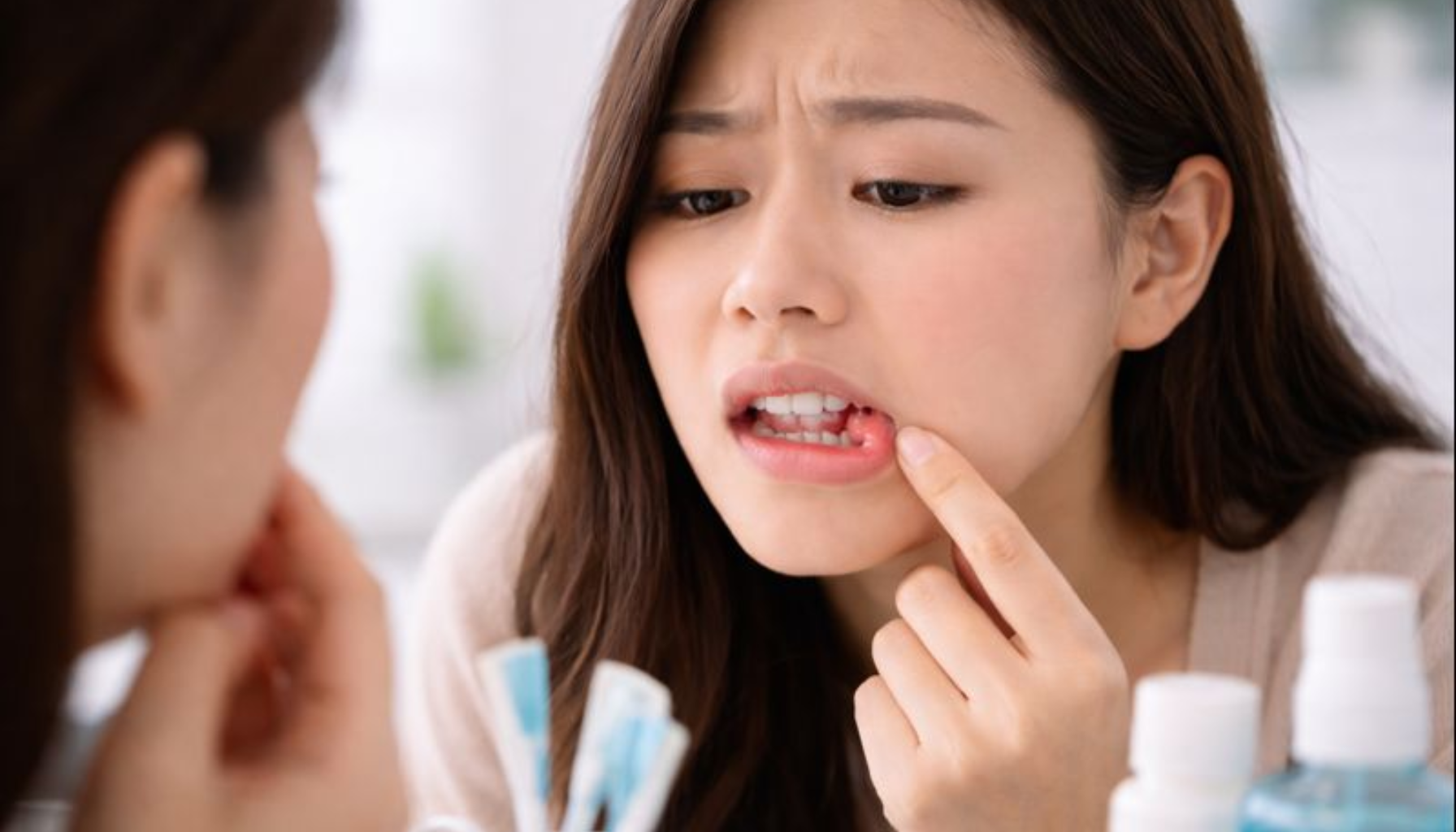 Blemme på Tannkjøttet: Når Små Hevelser Er Et Tegn på Irritasjon eller Infeksjon 17 Kvinne undersøker betent blemme på tannkjøttet foran speilet, omgitt av tannpleieprodukter på baderomsbenken.