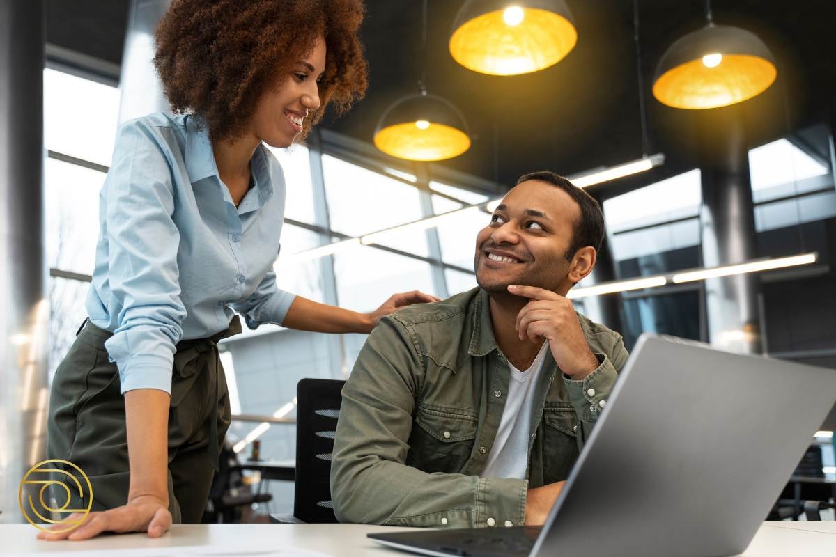 Friendly office interaction as a woman smiles while standing over a colleague seated at a desk, both engaged in conversation.