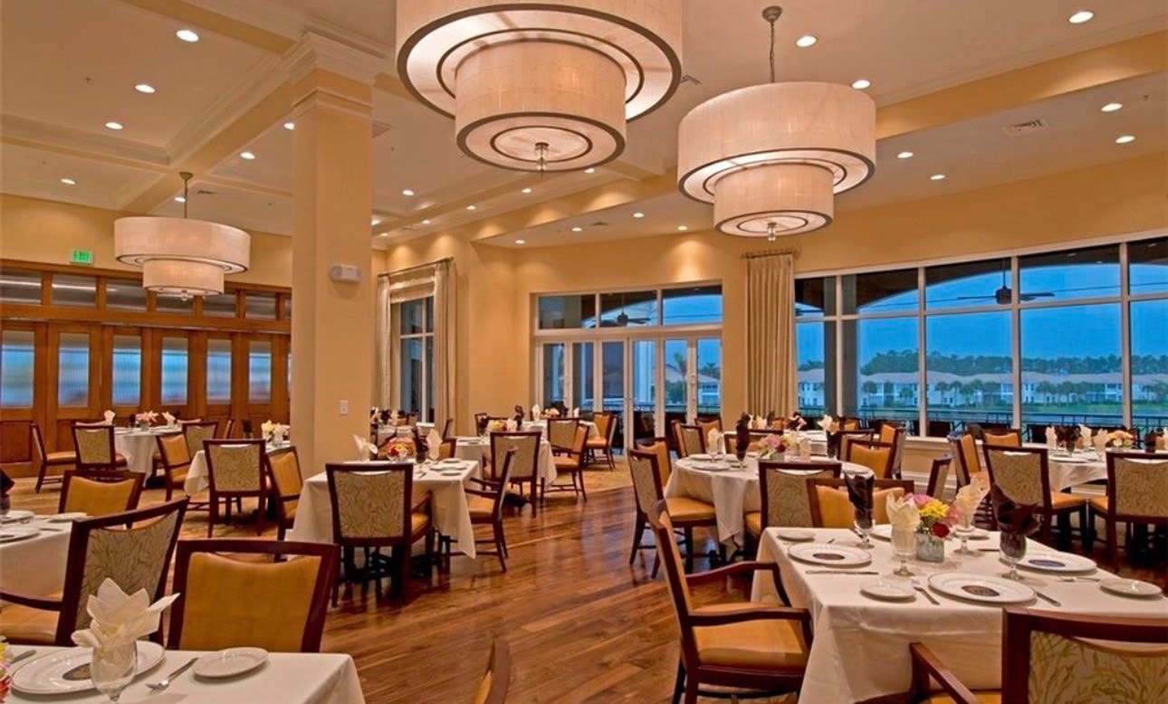 The image depicts an elegant clubhouse exterior at Naples Lakes Country Club, featuring a resort-style pool and tennis courts in the background, highlighting the community's luxurious amenities. This setting showcases the perfect blend of modern living and recreational opportunities for residents enjoying bundled golf membership.