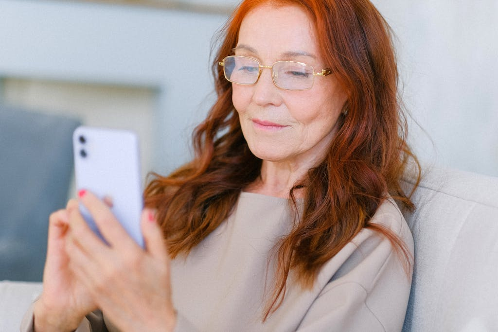 Mulher madura com cabelos ruivos usando óculos e olhando para seu smartphone em um ambiente interno.