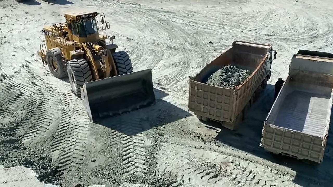 A large yellow Caterpillar loader with a full bucket preparing to dump its load into a dump truck, which already contains some material, with another empty dump truck parked nearby on a dirt construction site