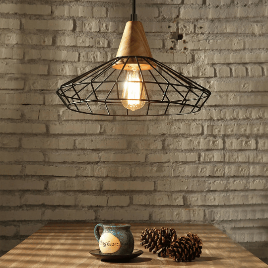 Rustic pendant light with a wooden top and black wire cage shade, hanging over a wooden table with a ceramic mug and pinecones, set against a white brick wall.