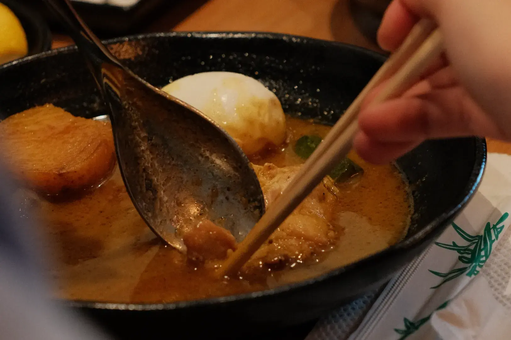 A person using chopsticks to retrieve food from a bowl with broth, containing pieces of meat and a soft-boiled egg.
