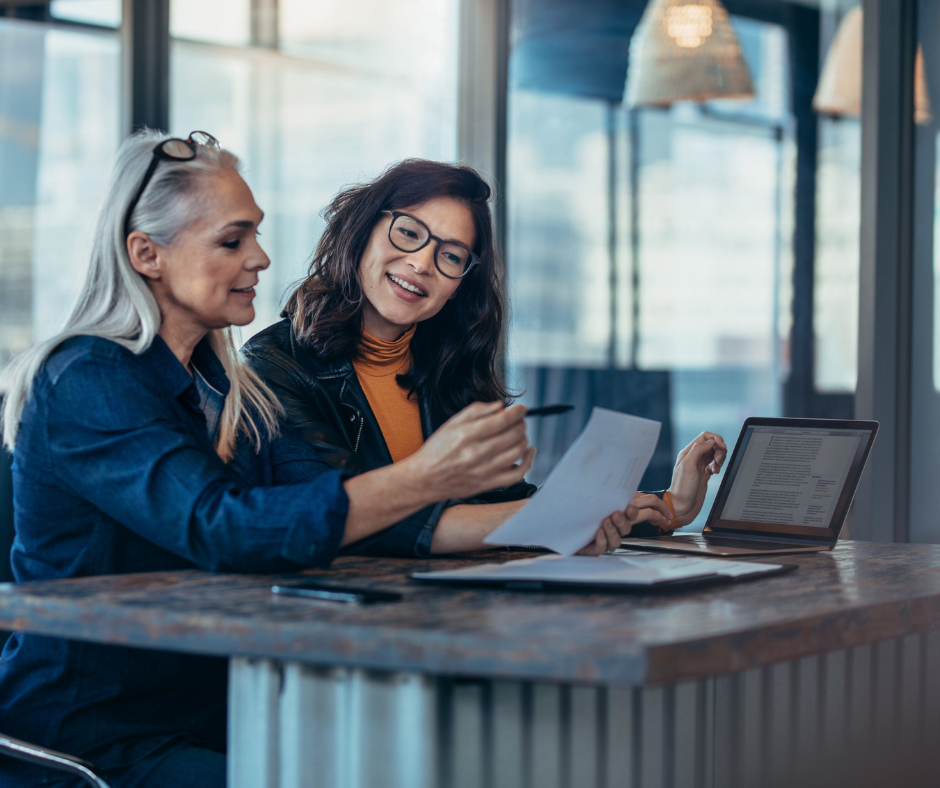 two women sit at a cafe table looking at a laptop and paper