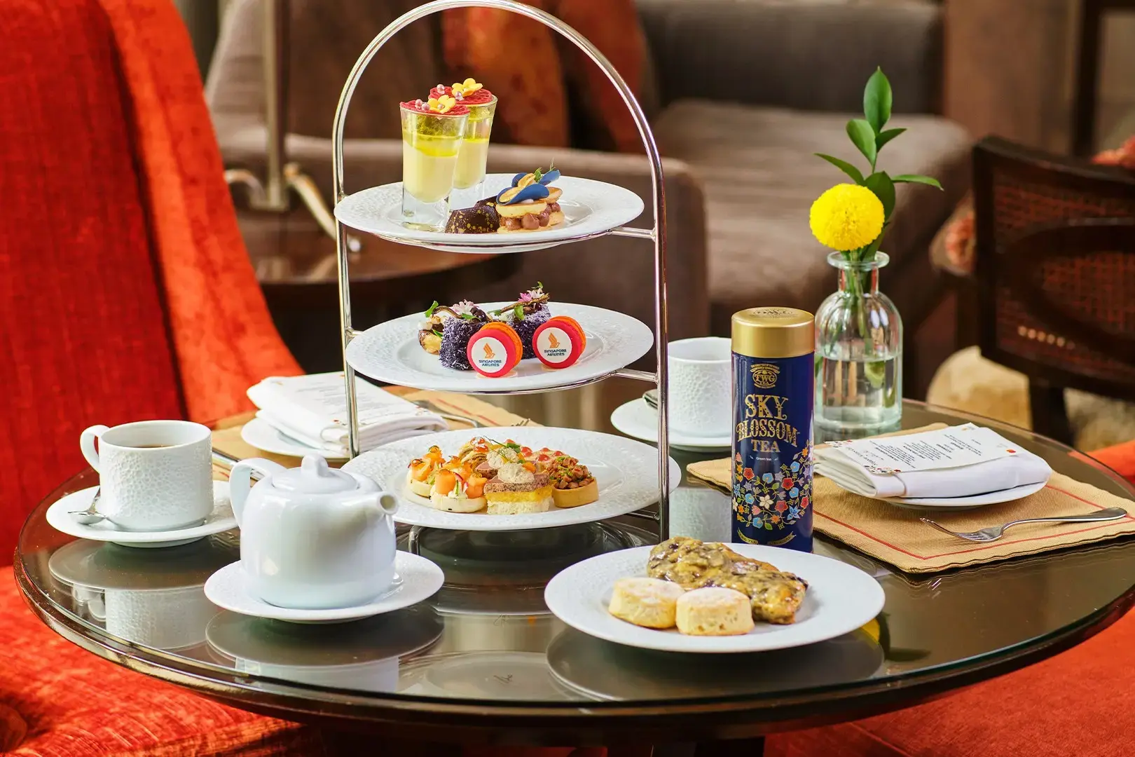 An elegant three-tiered afternoon tea set featuring delicate sweet and savory pastries is served on a glass table alongside a white teapot and cups. A blue and gold tin of "Sky Blossom Tea" and a single yellow flower in a vase complement the luxury spread, which includes branded macarons bearing the Singapore Airlines logo.