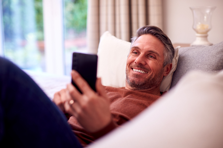 Gray haired man laying on a sofa checking his email.