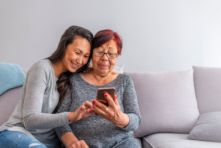 Older mom and adult daughter sitting on the sofa and smiling at something on a cell phone. 