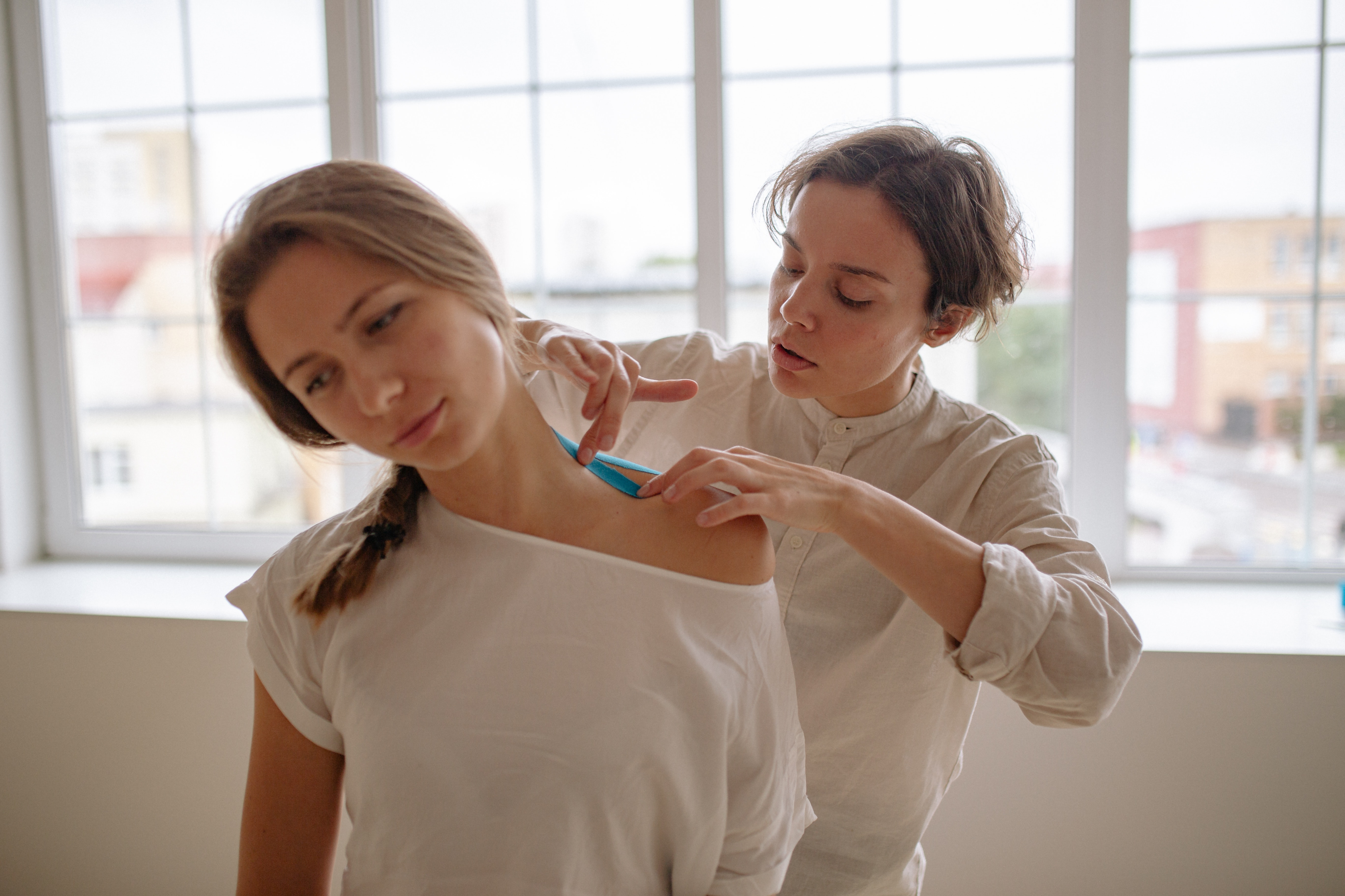 A woman applies ointment to another woman's neck, who appears uncomfortable, against a softly lit background from a window.