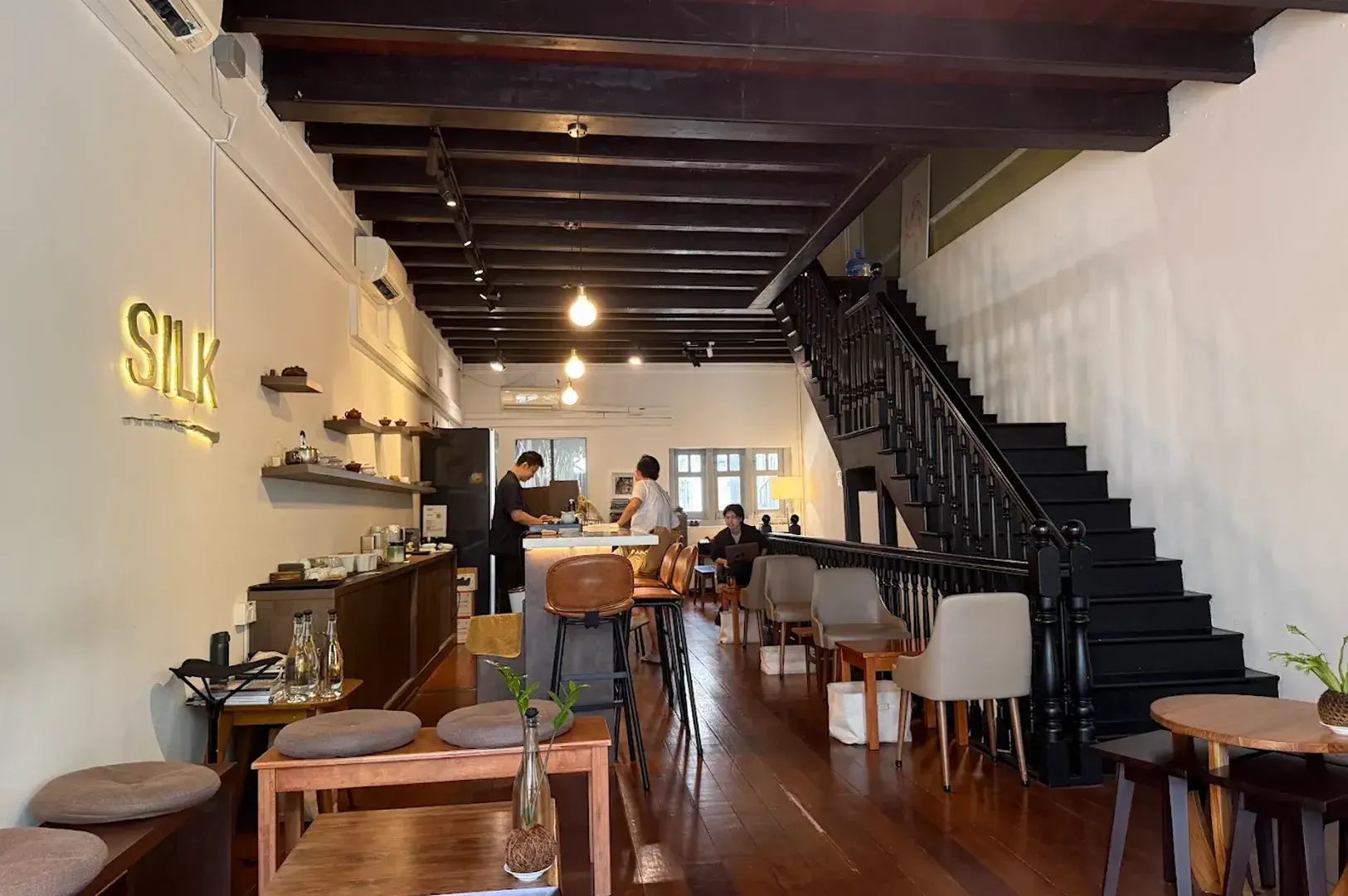 Wide view of a modern shophouse tea lounge with exposed wooden ceiling beams, a grand staircase, and an illuminated "SILK" sign.