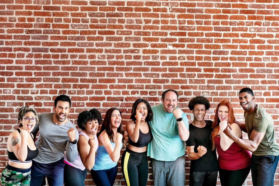 A diverse group of nine people standing against a brick wall, smiling and posing with fists raised in excitement and determination.