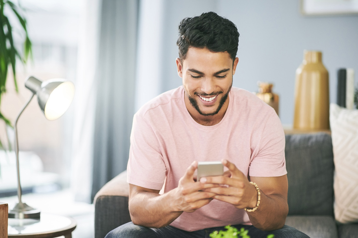 Cheerful young man in a pink tee shirt sitting on a sofa and looking at his smartphone.