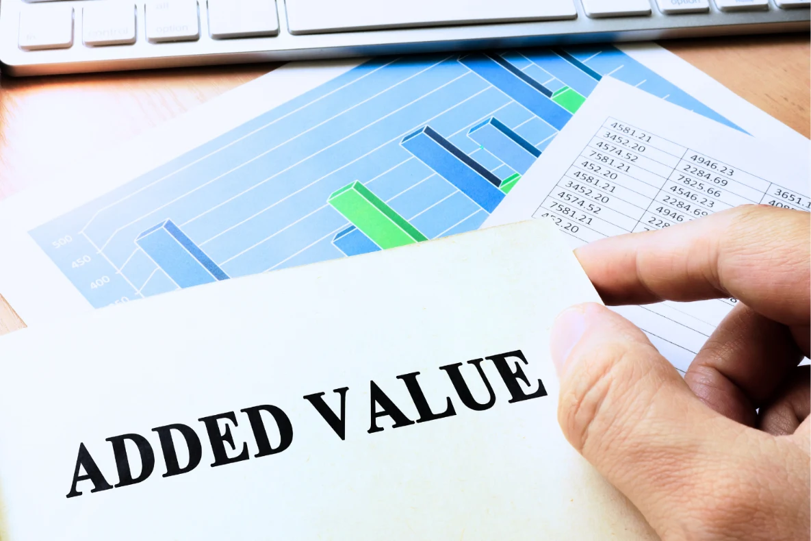 A hand holds a document with the words ADDED VALUE in large black letters, over charts and financial data on a desk with a keyboard and mouse.