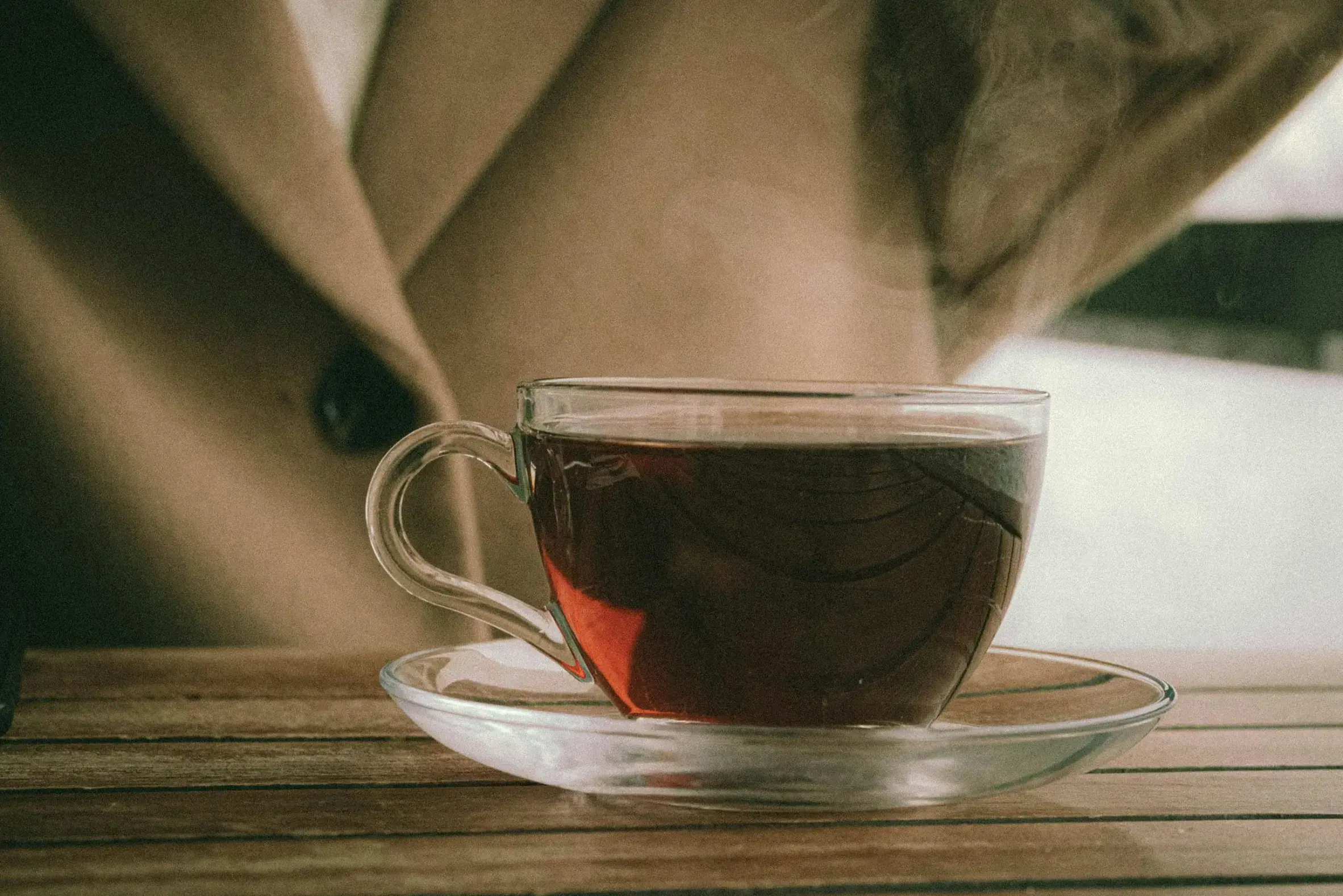 A steaming glass cup filled with dark amber tea sits on a matching clear saucer on a wooden table. In the soft-focus background, a person wears a cozy tan or beige winter coat with a large black button.