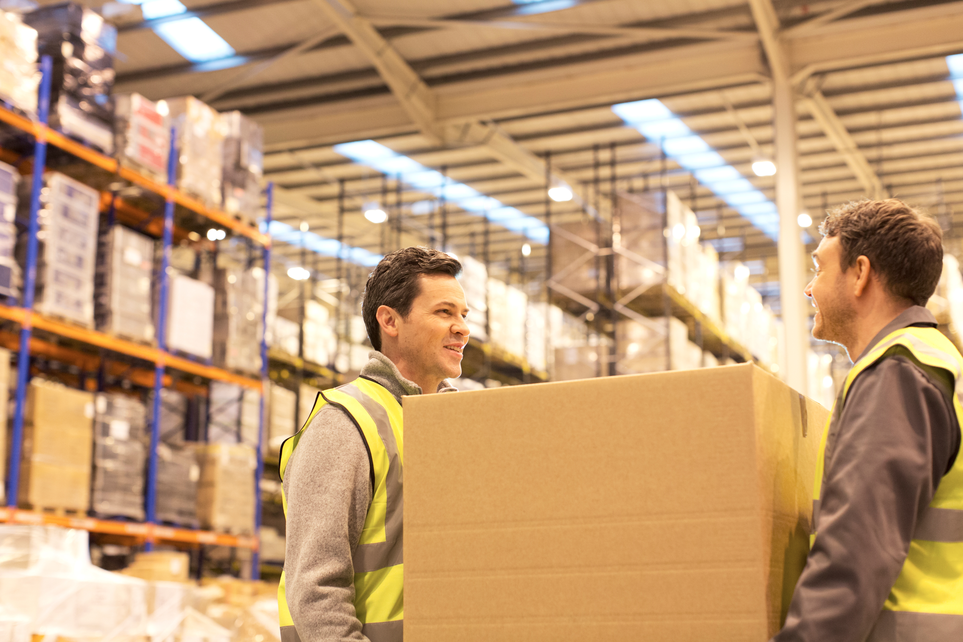 Two men wearing yellow vests carry a box in a warehouse.