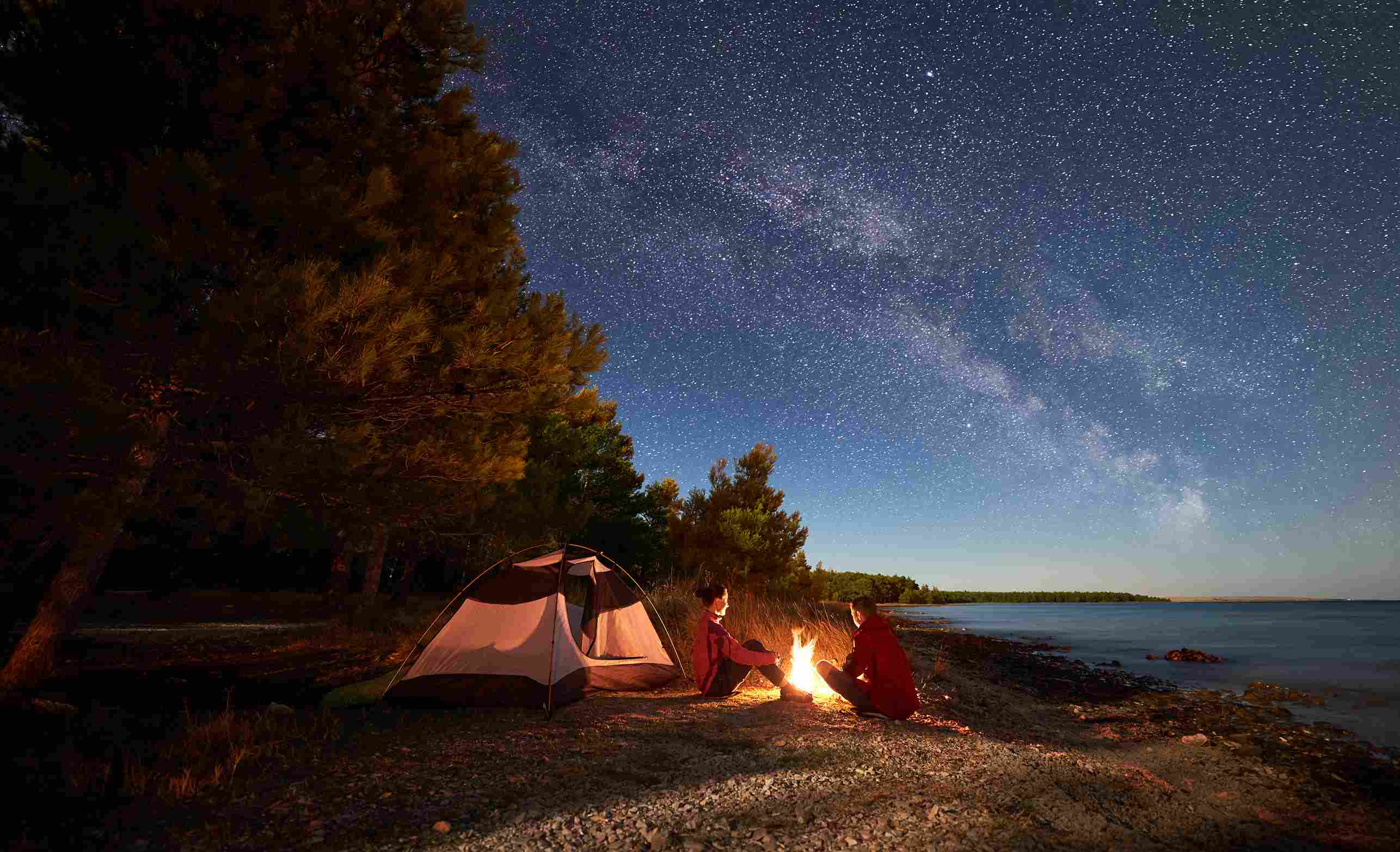 Dwie osoby siedzące przy ognisku przed namiotem na brzegu jeziora w nocy