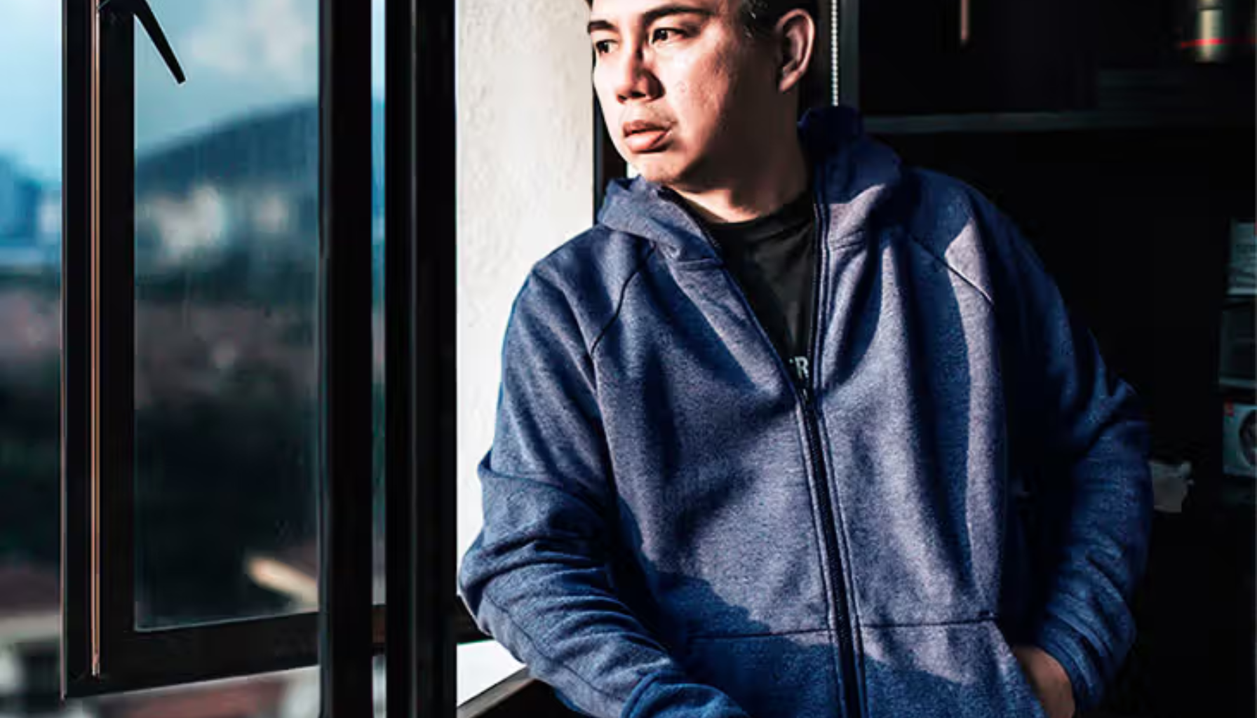 Mann i blå hettegenser står ved åpent vindu i sollys og puster rolig.