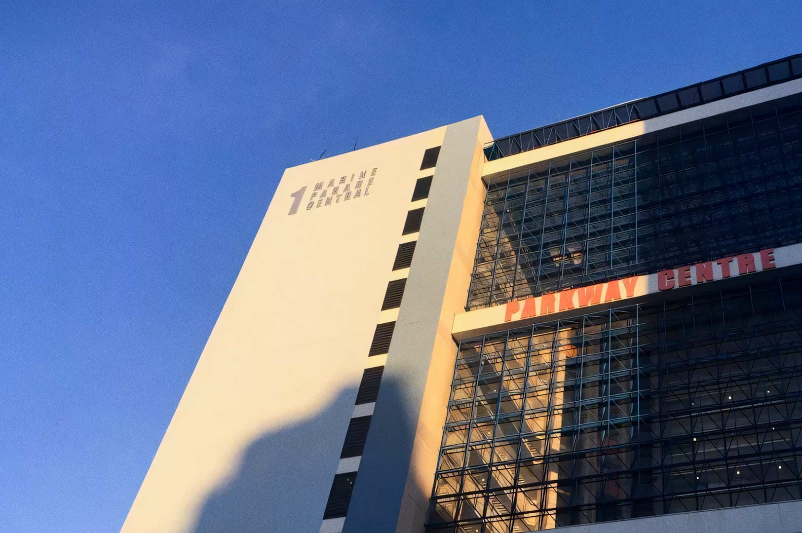 Low-angle view of the Parkway Centre building showing its tall white façade and adjacent glass-fronted section with the words ‘PARKWAY CENTRE’ visible. The building is lit by warm sunlight against a clear blue sky.