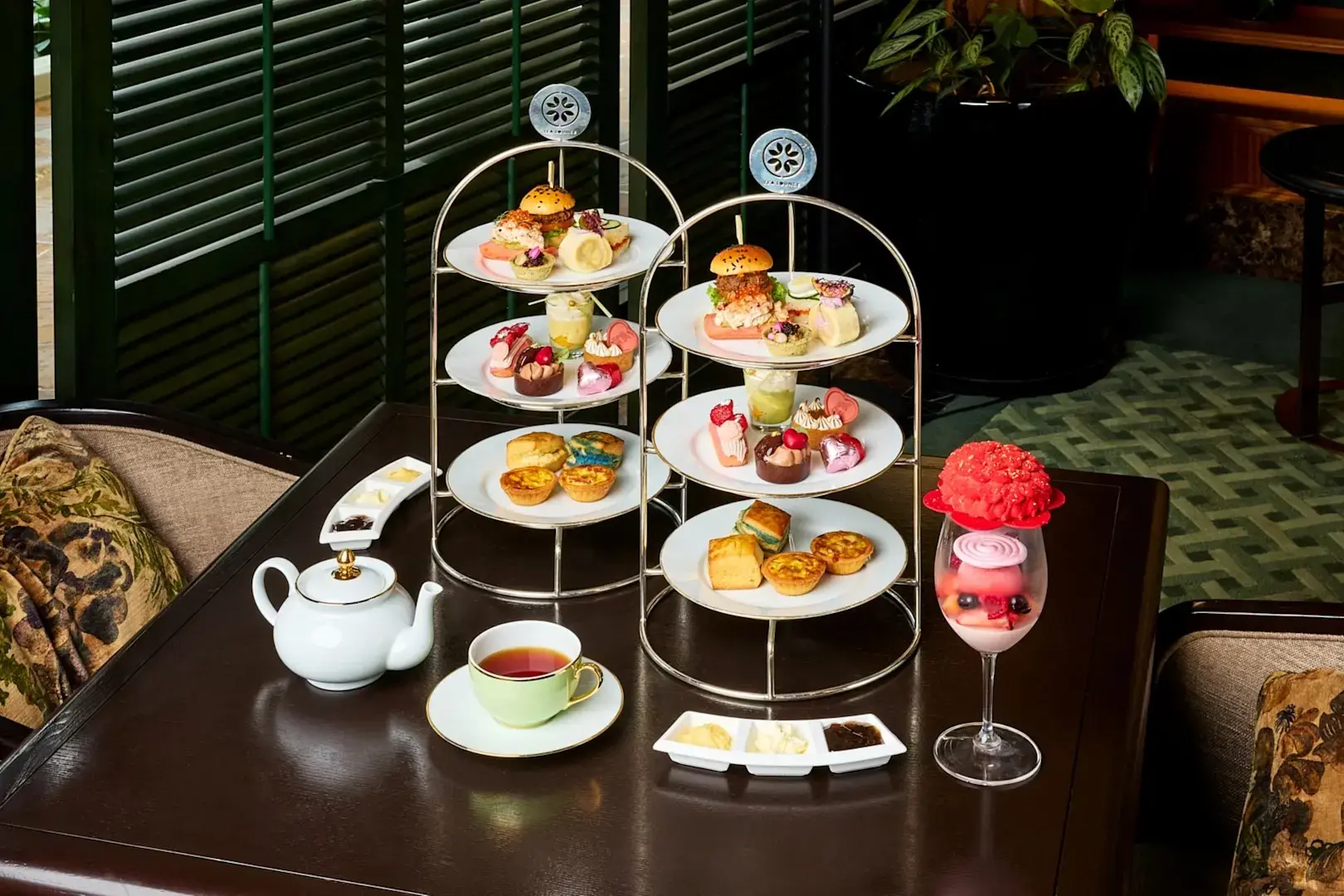 An elegant afternoon tea set is displayed on a dark wooden table, featuring two tiered stands filled with colorful savory sliders, delicate pastries, and warm scones. A white teapot, a cup of tea, and a decorative glass dessert topped with a vibrant red crumble complete the sophisticated arrangement.