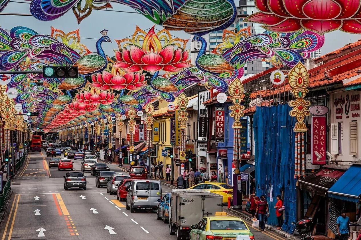 This vibrant street scene captures the bustling atmosphere of Little India in Singapore, adorned with elaborate overhead decorations featuring peacocks and lotus flowers. The busy road is lined with colorful shophouses and traditional signage, showcasing a mix of local heritage and modern urban life.