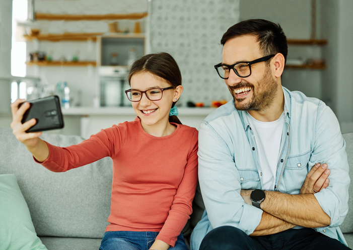 Cute dad and daughter, both in glasses, watching something on her cell and laughing.