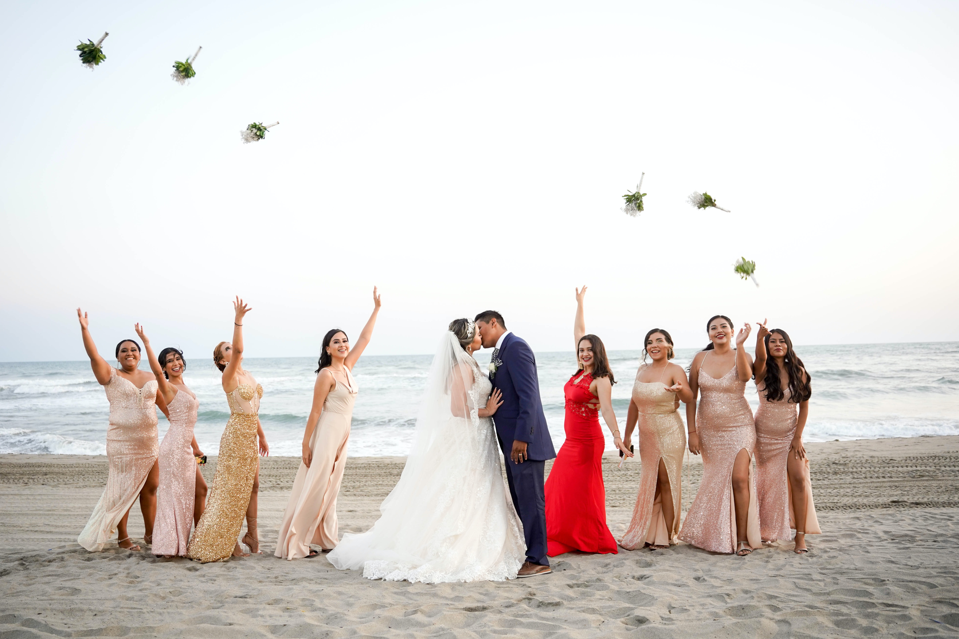 The couple is kissing on the beach, surrounded by women tossing their flowers.