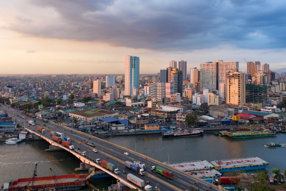 Urban skyline of Metro Manila with high-rise buildings and a river bridge during sunset.