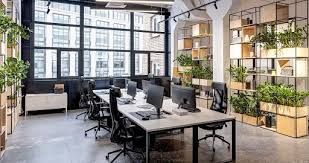 A modern office space with large windows providing ample natural light. The office features several desks with desktop computers and ergonomic chairs arranged in a collaborative setup. There are numerous plants throughout the space, both on shelves and around the desks, creating a fresh and inviting atmosphere. The design includes clean lines, a neutral color palette, and open shelving, enhancing the open and airy feel of the workspace.