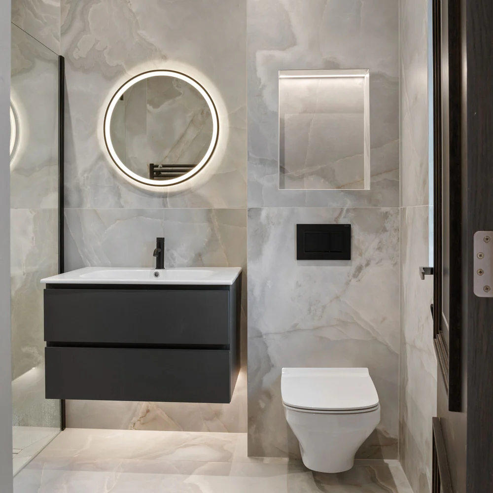 Modern bathroom with a floating vanity, round backlit mirror, and wall-mounted toilet against marble-effect tiles.