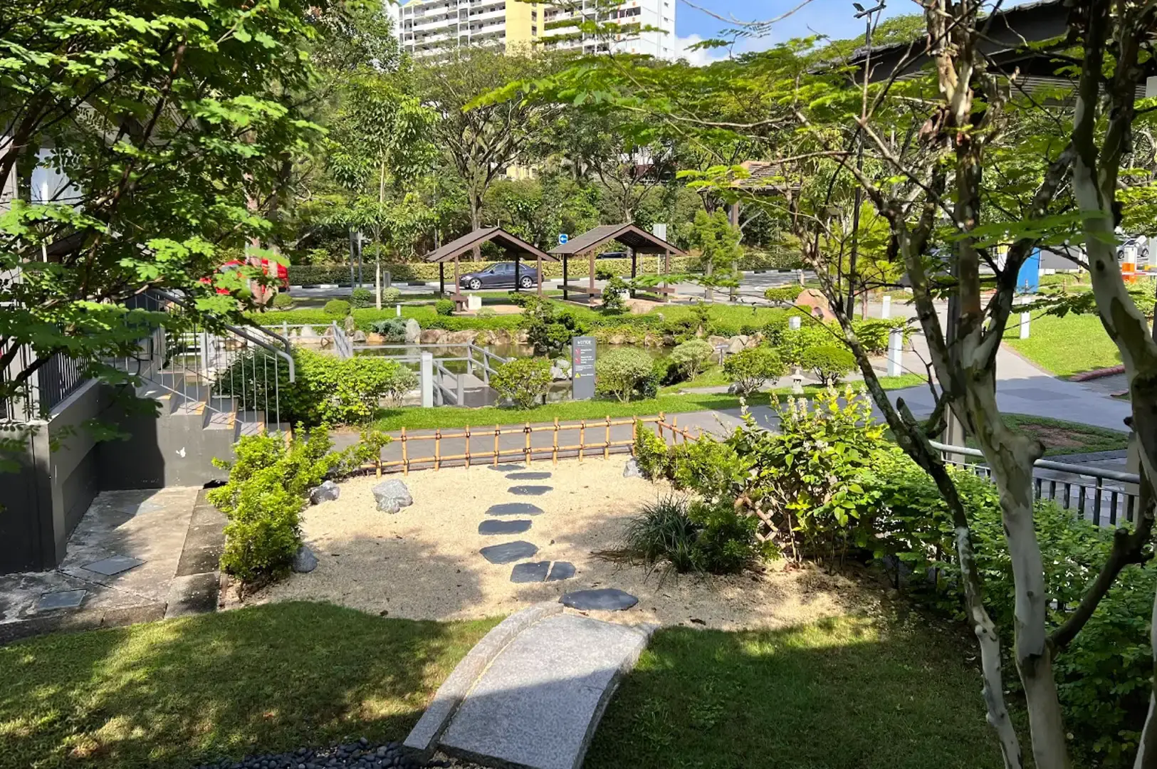 A elevated view framed by green leafy branches showing a stone stepping-stone path leading across raked gravel and grass patches toward the pond area and wooden gazebos, with an outdoor staircase leading down from the restaurant building on the left.