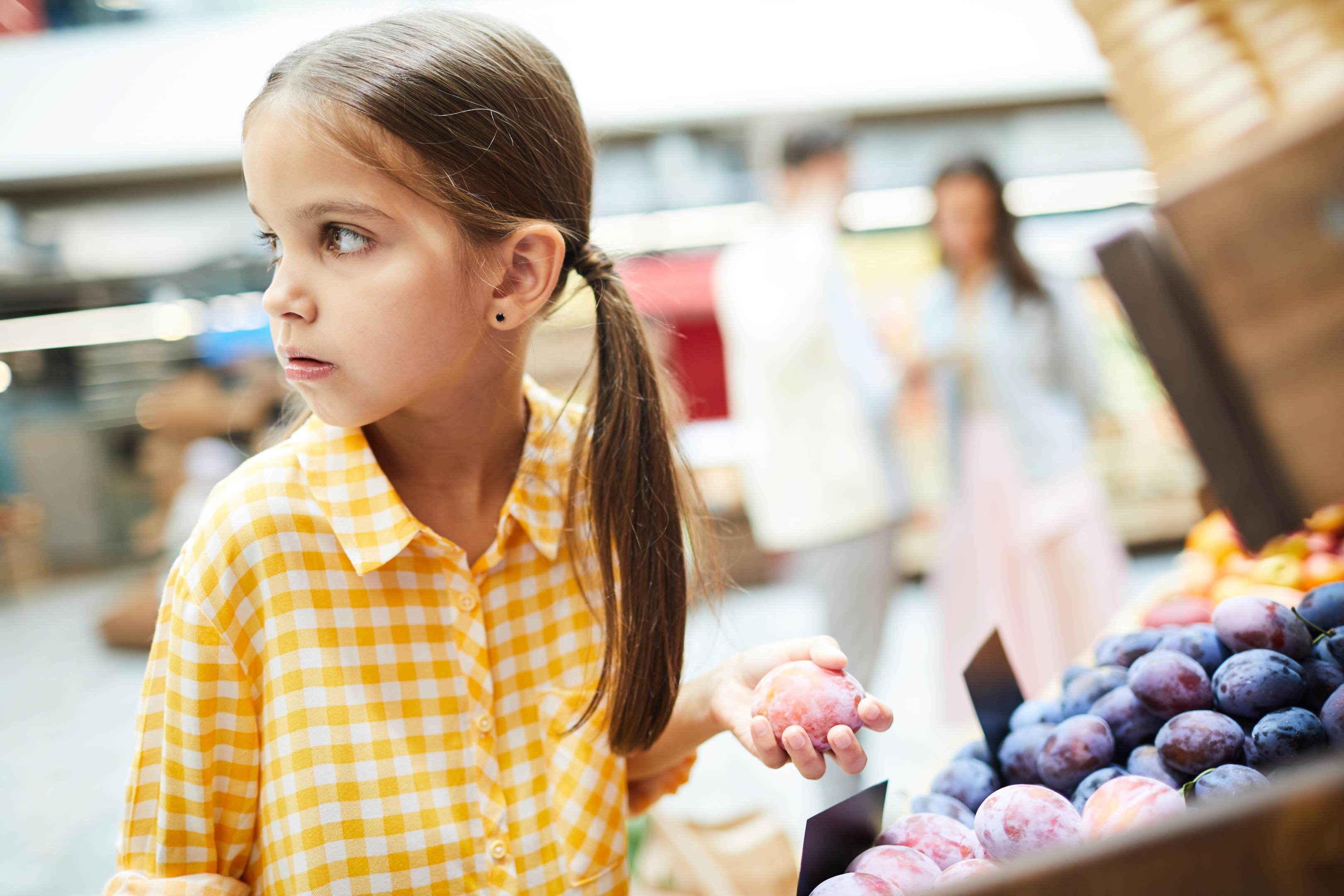 Concerned girl with ponytails, dressed in a checkered shirt, stands by the food shelves holding a plum, contemplating the consequences and deciding against stealing it in the grocery store.