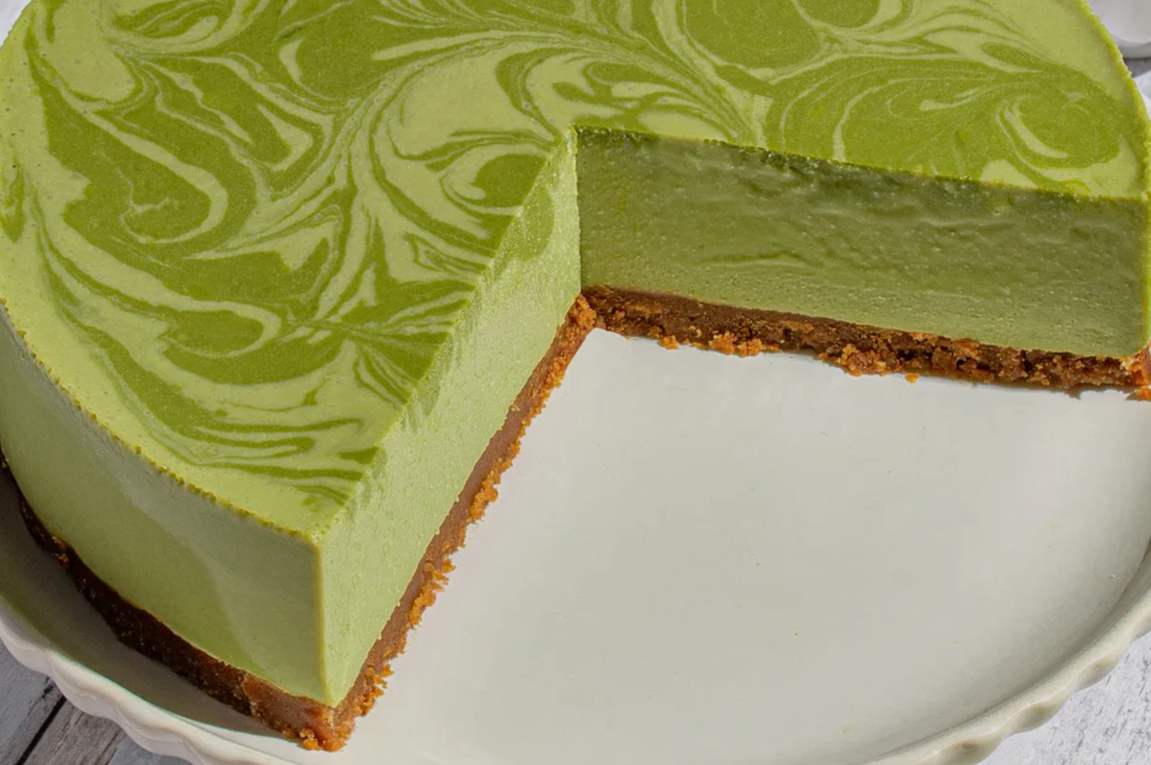 A round matcha cheesecake with a marbled green top and a slice missing, revealing a smooth texture and a brown crust, on a white plate.