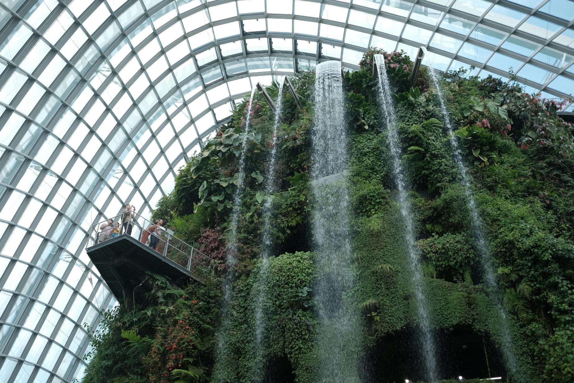 Inside a massive glass and steel dome, a spectacular man-made mountain covered in lush vegetation features a tall waterfall cascading down its side. Visitors stand on a suspended walkway to admire the mist and the intricate architectural grid of the curved roof above.