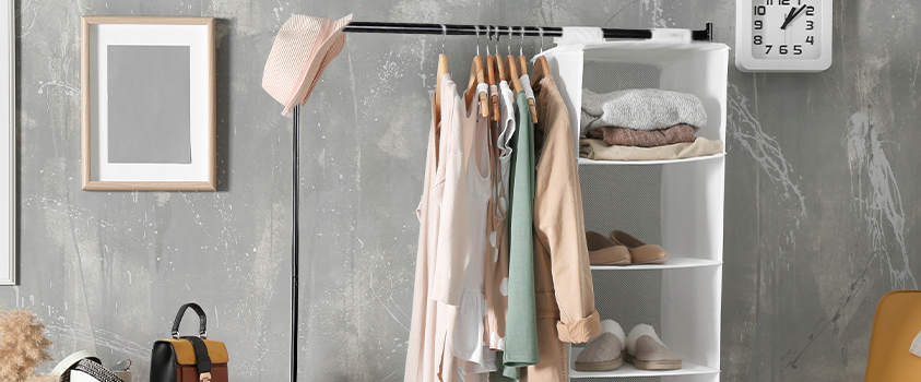An Artiss 6ft Black Metal Garment Rack holding a variety of neutral coloured clothes and a white hanging storage unit. The unit is filled with shoes and folded shirts. It is in a bedroom with a grey concrete wall. 